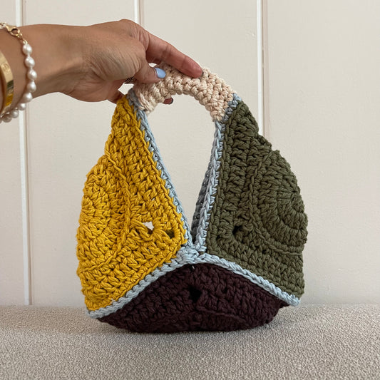 A hand holding a crocheted grannhy square handbag in yellow, green, brown, baby blue, and cream. 
