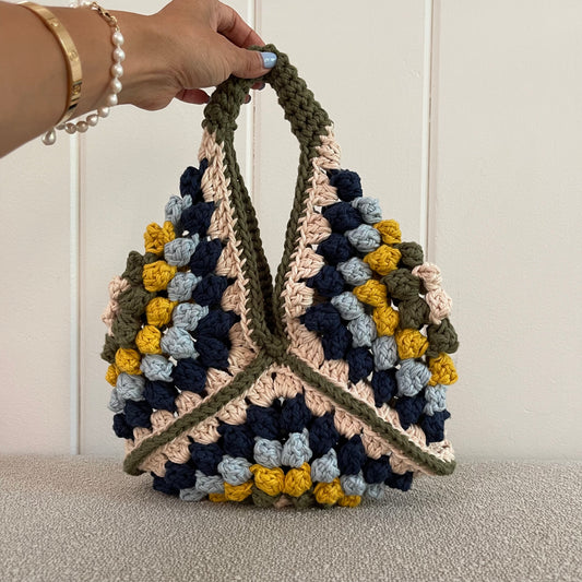 a hand holding a crocheted handbag in cream, green, yellow, baby blue and navy blue. 